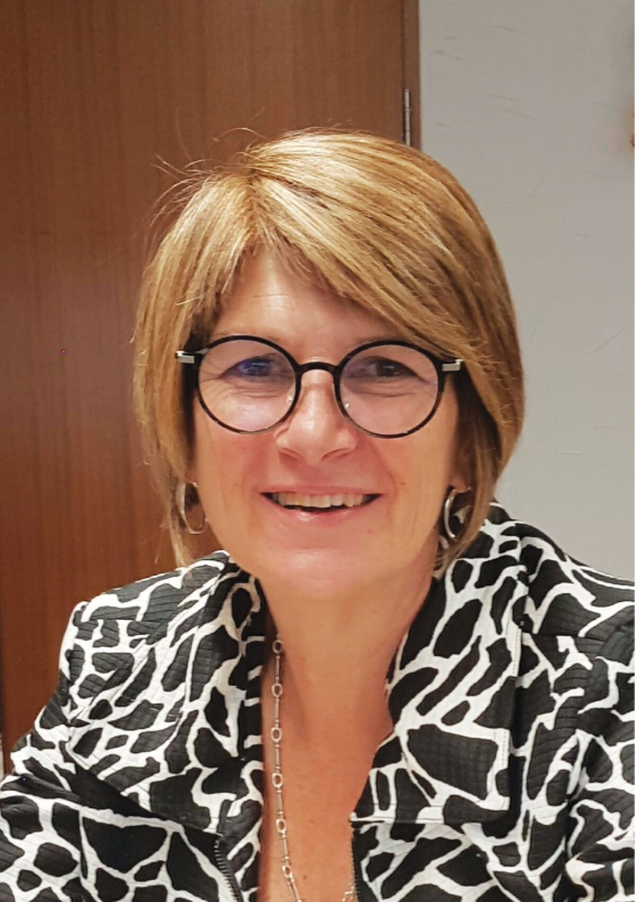 Portrait of Dr Frances Hughes seated indoors, wearing a patterned black‑and‑white jacket and a necklace, photographed in an office setting with a wooden door and light‑coloured wall in the background.