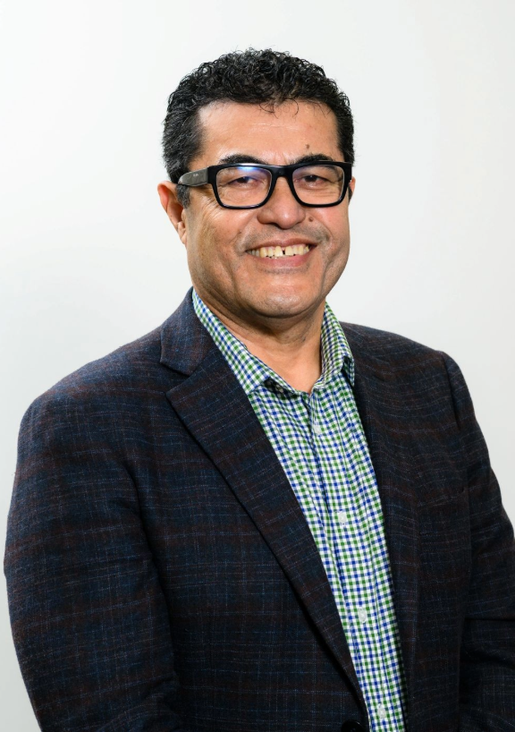 Portrait of Iosefa Tiata Paituli wearing a dark blazer over a green‑and‑blue checked shirt, photographed against a plain light background.