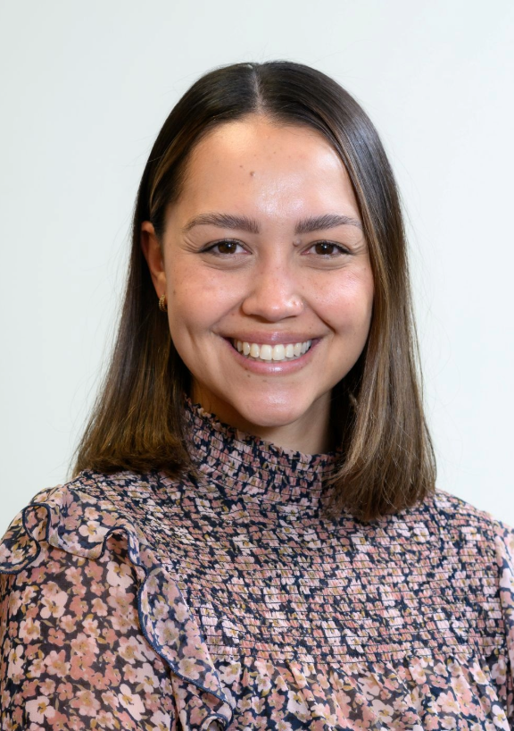 Portrait of Miriam Manga wearing a high‑neck patterned blouse, photographed against a plain light background.