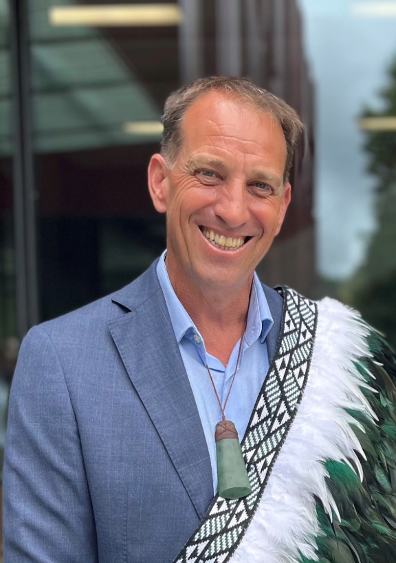 Portrait of Tony Ward standing outdoors near a modern glass building, wearing a light blue jacket and shirt with a traditional korowai draped over one shoulder and a carved pendant necklace.