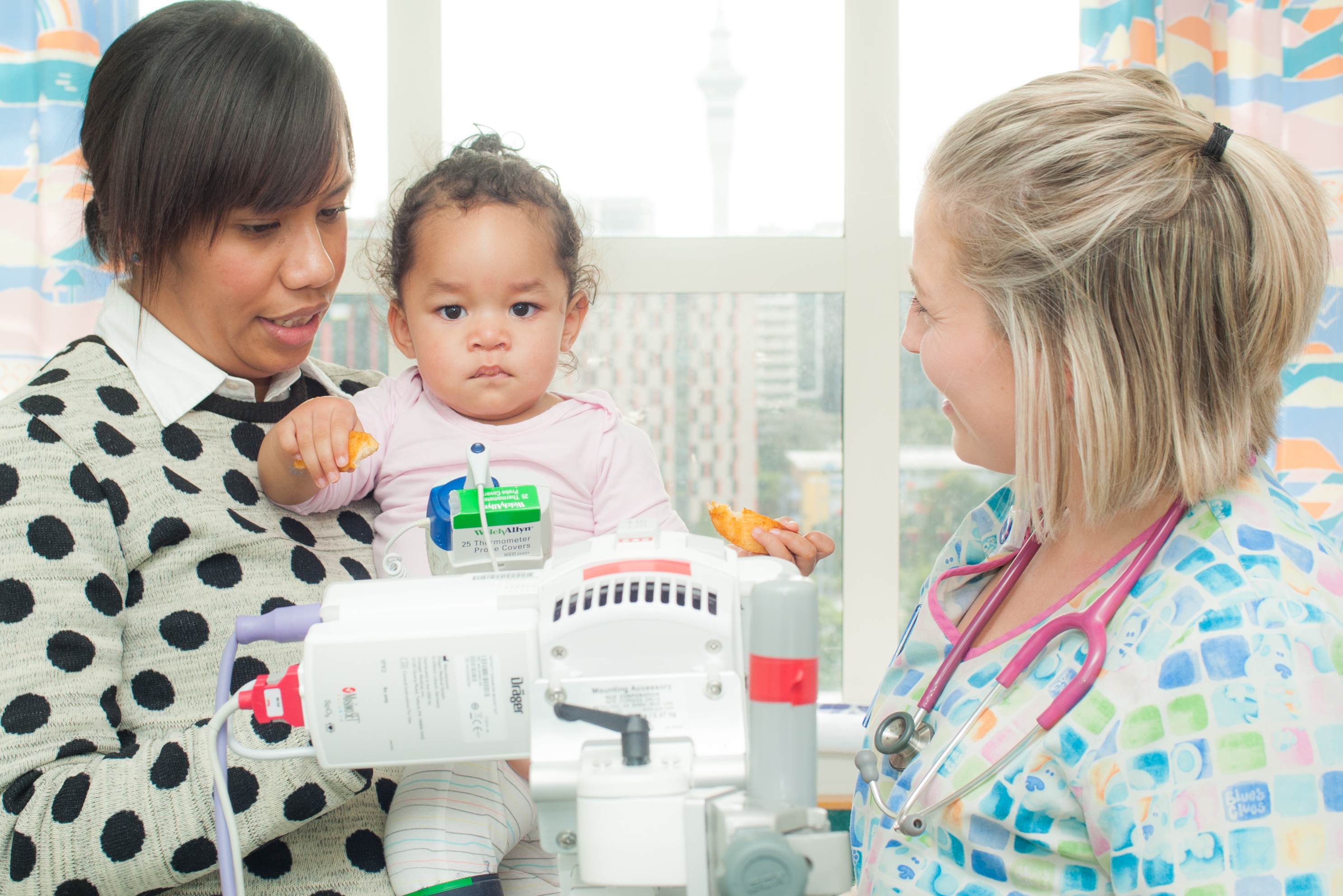 An adult caregiver holding a young child beside a hospital bed, with a nurse wearing a stethoscope standing nearby. Medical monitoring equipment is positioned in front of the child, and a hospital window with a city view is visible in the background.