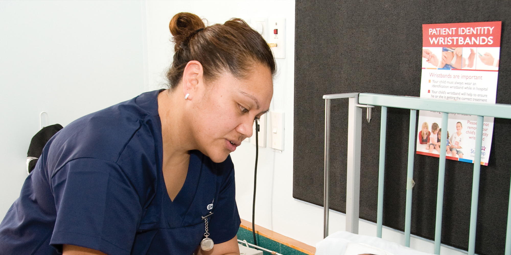 A nurse in clinical scrubs leans over a hospital cot, providing care in a ward setting. A wall notice about patient identity wristbands and clinical furnishings are visible nearby, indicating a healthcare environment.